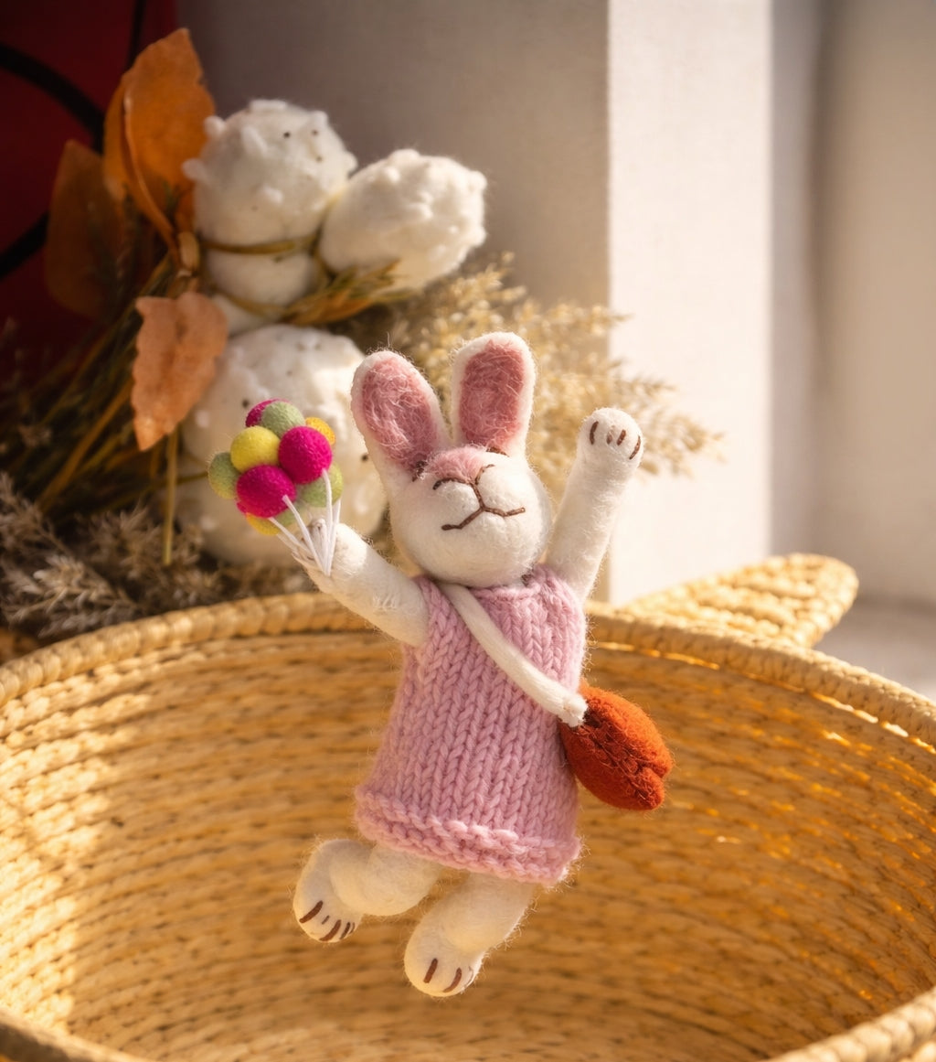 Plush toy rabbit in a pink dress holding balloons, placed on a woven surface with decorative elements in the background.