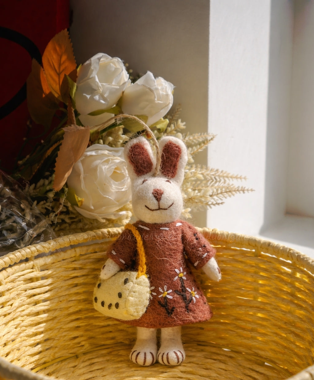 Small rabbit wool felt in a brown dress holding a yellow bag, surrounded by white flowers and leaves on a woven surface.
